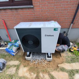 Michał Bieroń - Instalacja pompy ciepła Vaillant aroTHERM plus na betonowych fundamentach z podsypką żwirową, obok narzędzia i worki z kamieniami, widoczny fragment muru z cegły.