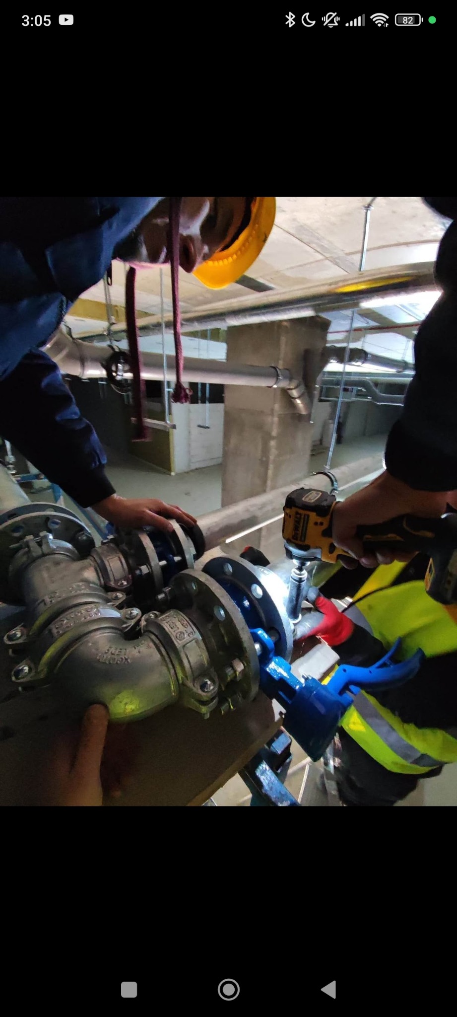 Instalator w kasku dokręca śruby kołnierza rurociągu za pomocą wkrętarki akumulatorowej DeWalt, widoczne rury i zawory w pomieszczeniu technicznym.