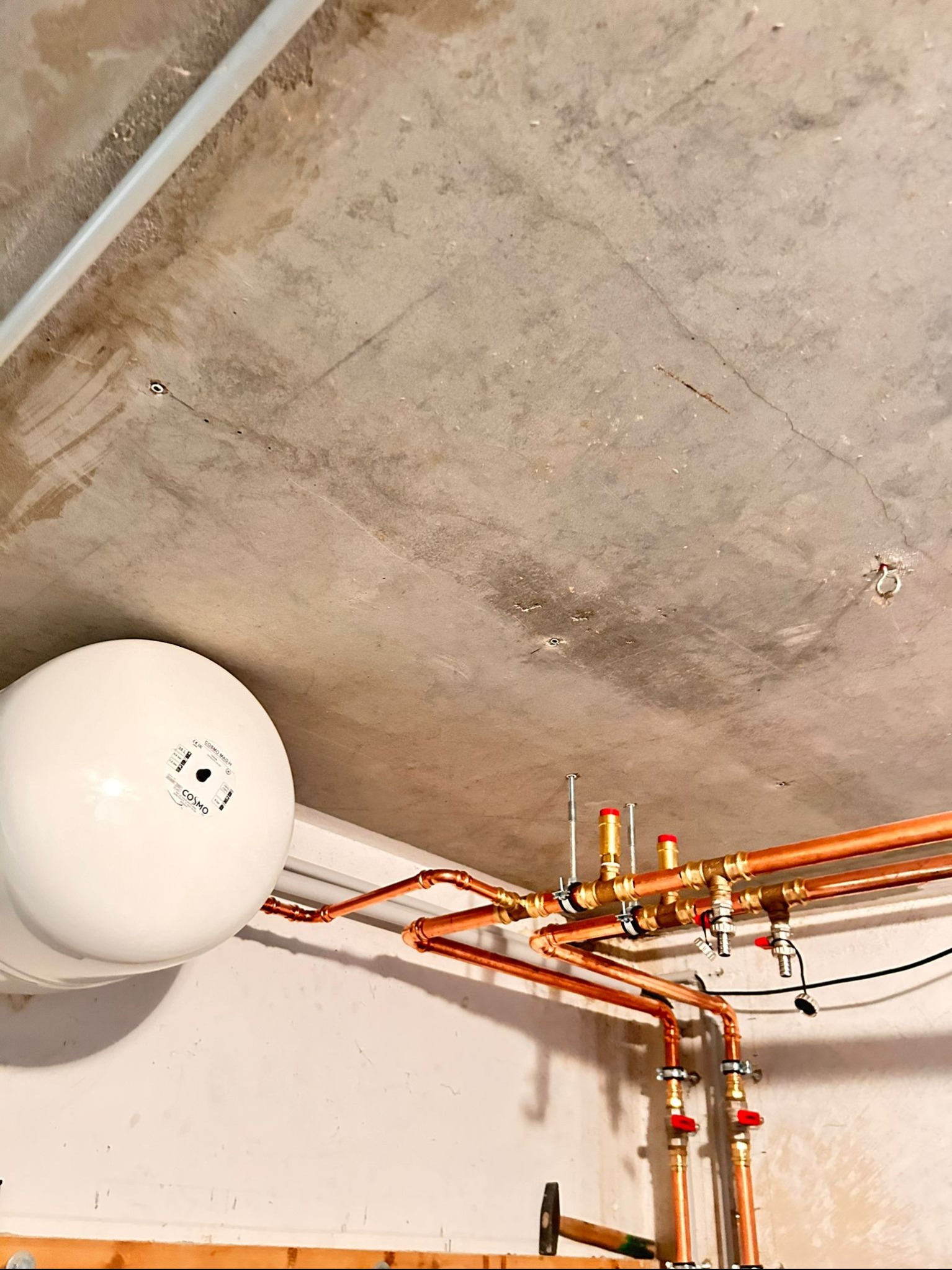 Biały zbiornik wyrównawczy CO, miedziane rury instalacji grzewczej z zaworami, widoczne pod betonowym sufitem, młotek w tle.