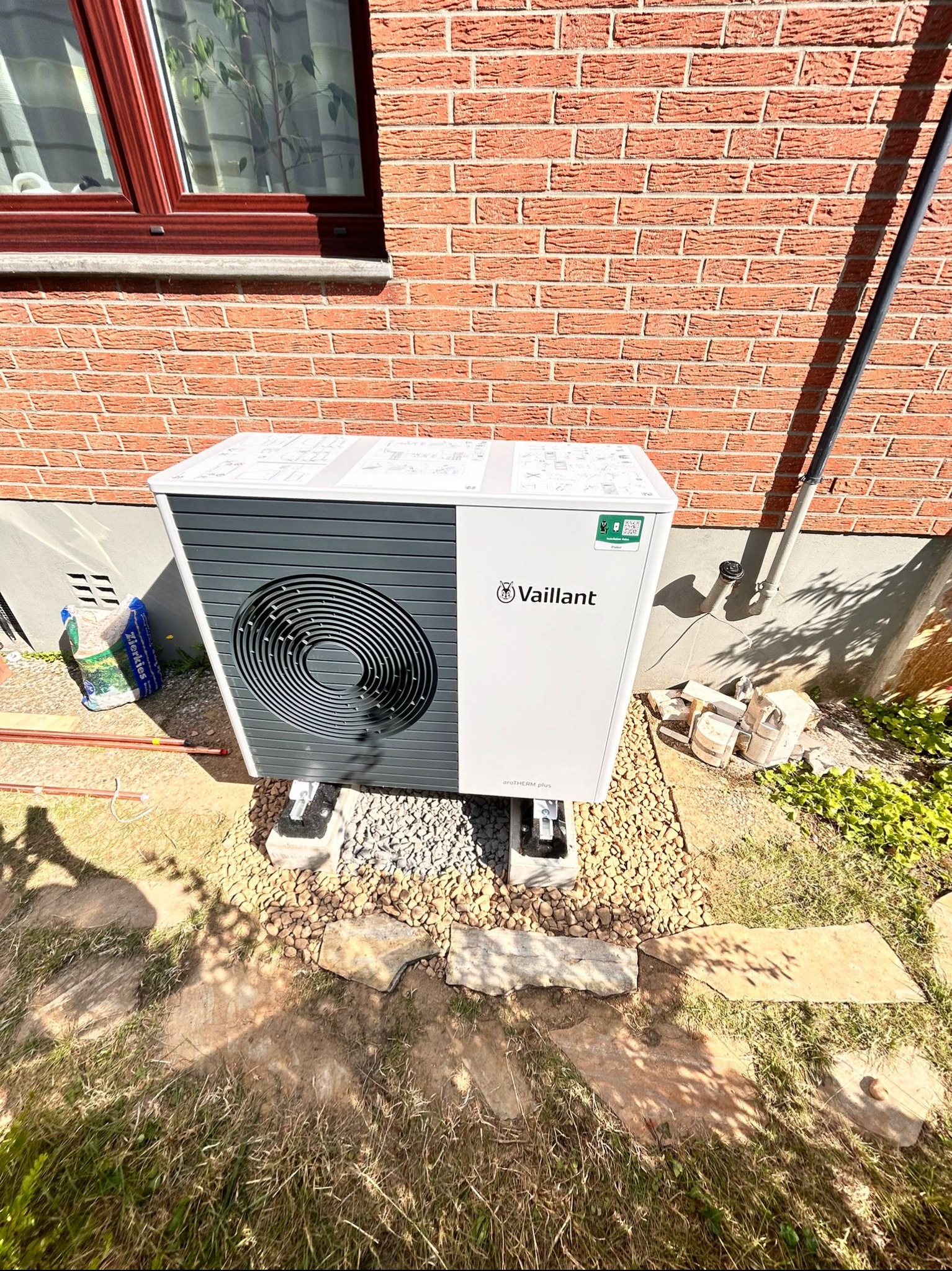 Pompa ciepła Vaillant zamontowana na betonowych bloczkach i żwirowym podłożu przy ceglanej ścianie budynku z czerwonym oknem, widoczne rury miedziane i worek z kamykami.