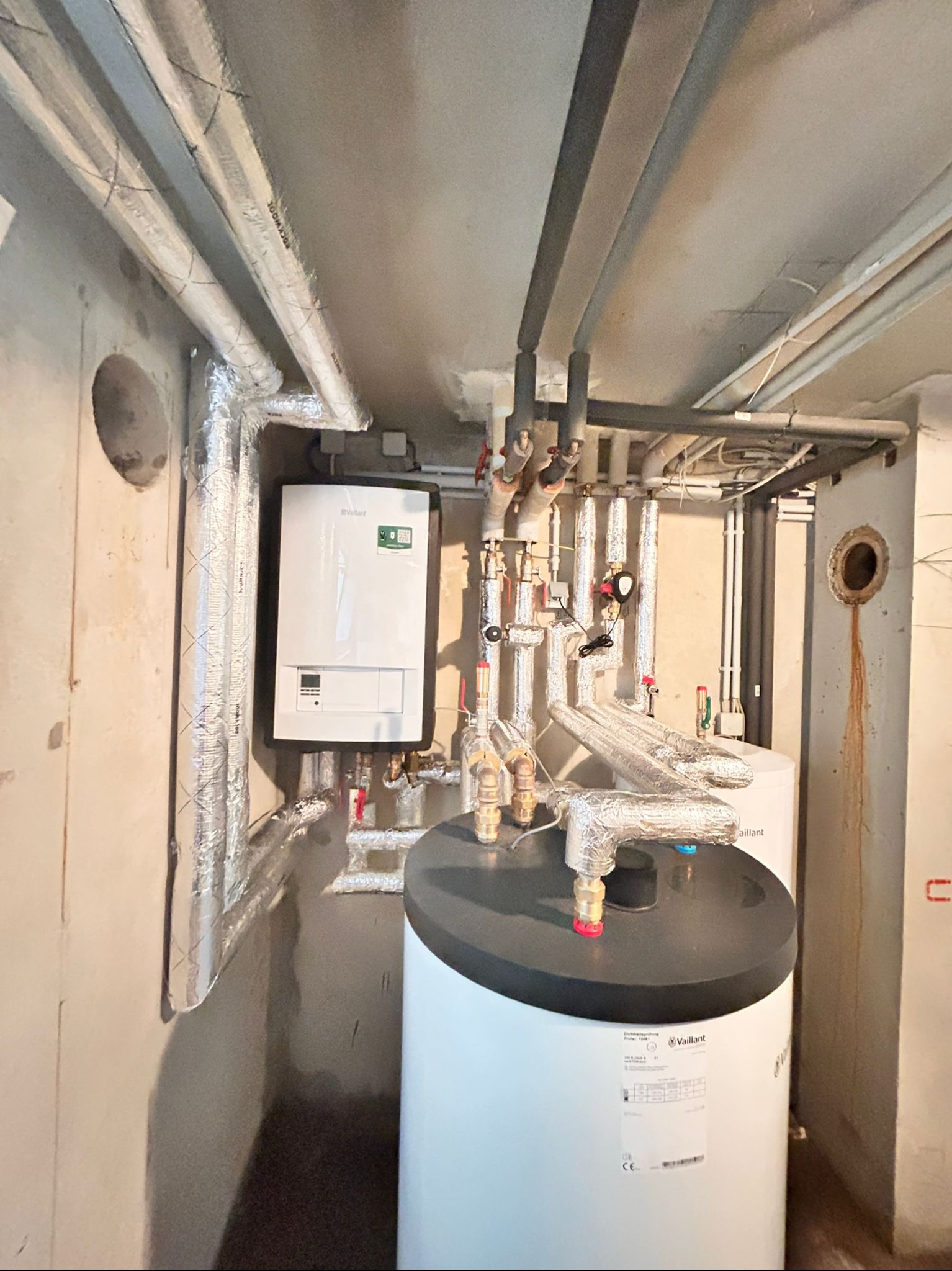 Kotłownia w piwnicy: biały piec gazowy Vaillant z czarną obudową, podłączony do instalacji z izolowanymi rurami miedzianymi, widoczne elementy armatury, na ścianie ślady po wywierconym otworze.
