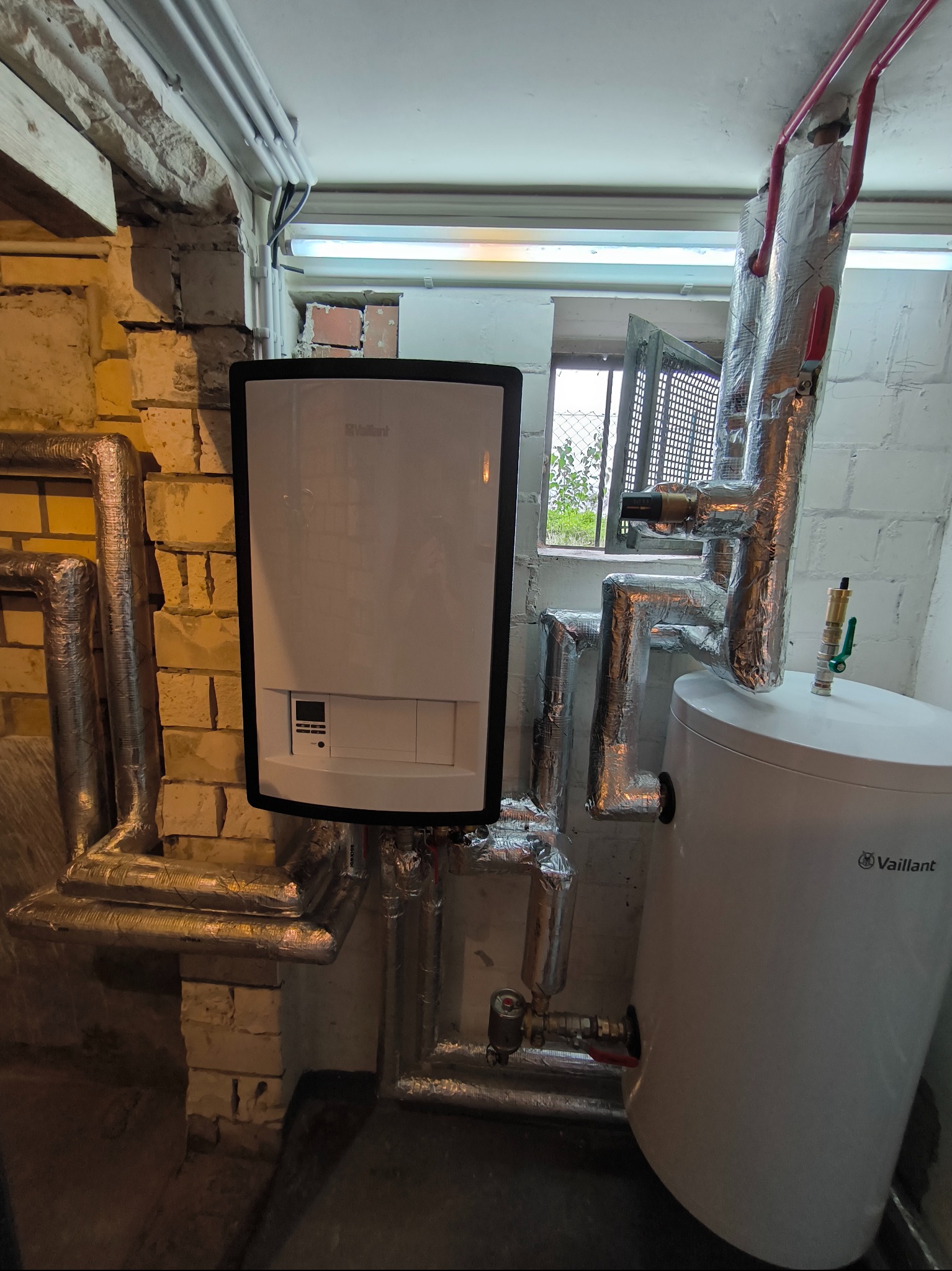 Kocioł gazowy Vaillant zamontowany w piwnicy, z widocznymi instalacjami i oknem z kratą w tle.