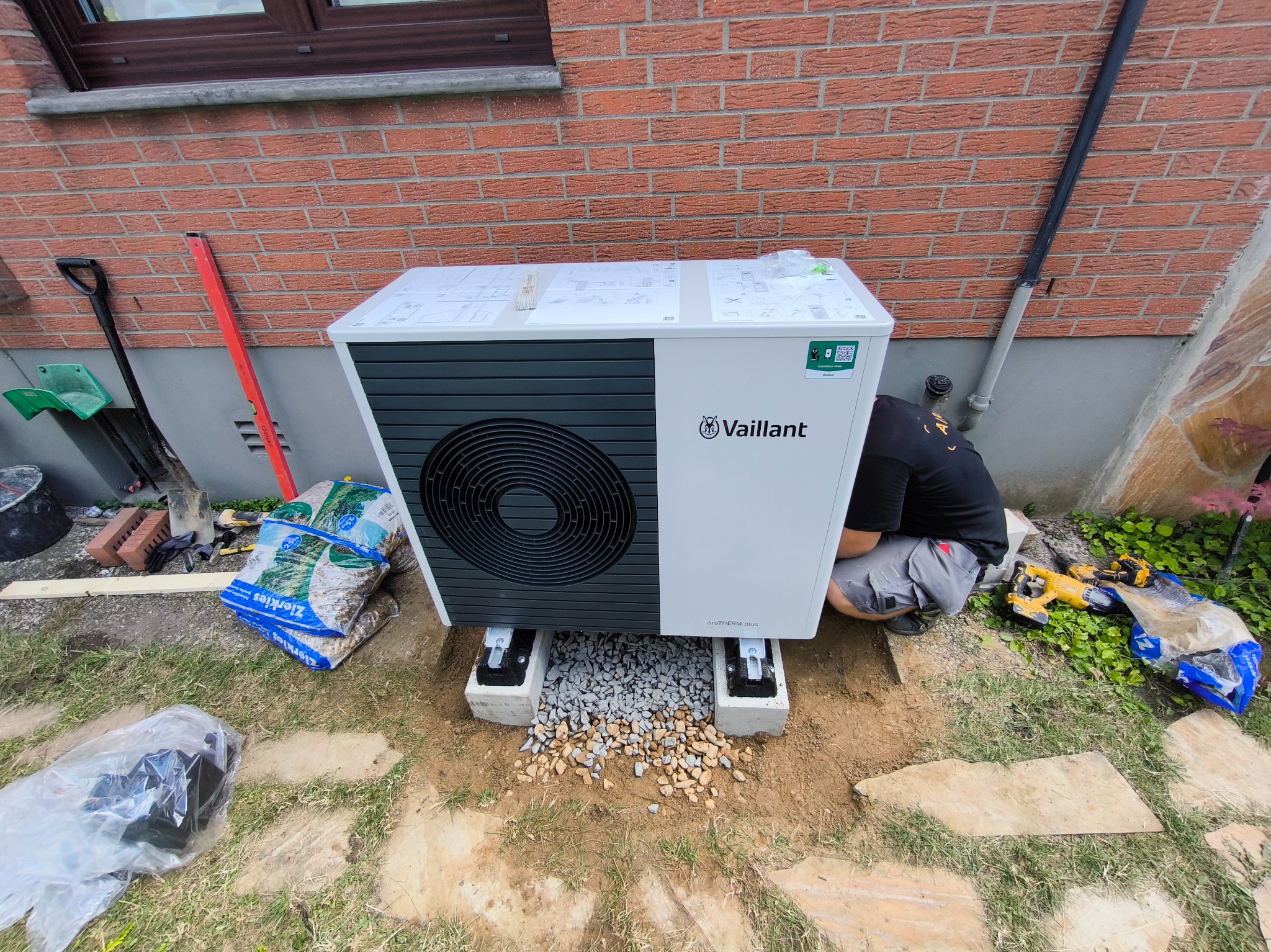 Instalacja pompy ciepła Vaillant aroTHERM plus na betonowych fundamentach z podsypką żwirową, obok narzędzia i worki z kamieniami, widoczny fragment muru z cegły.