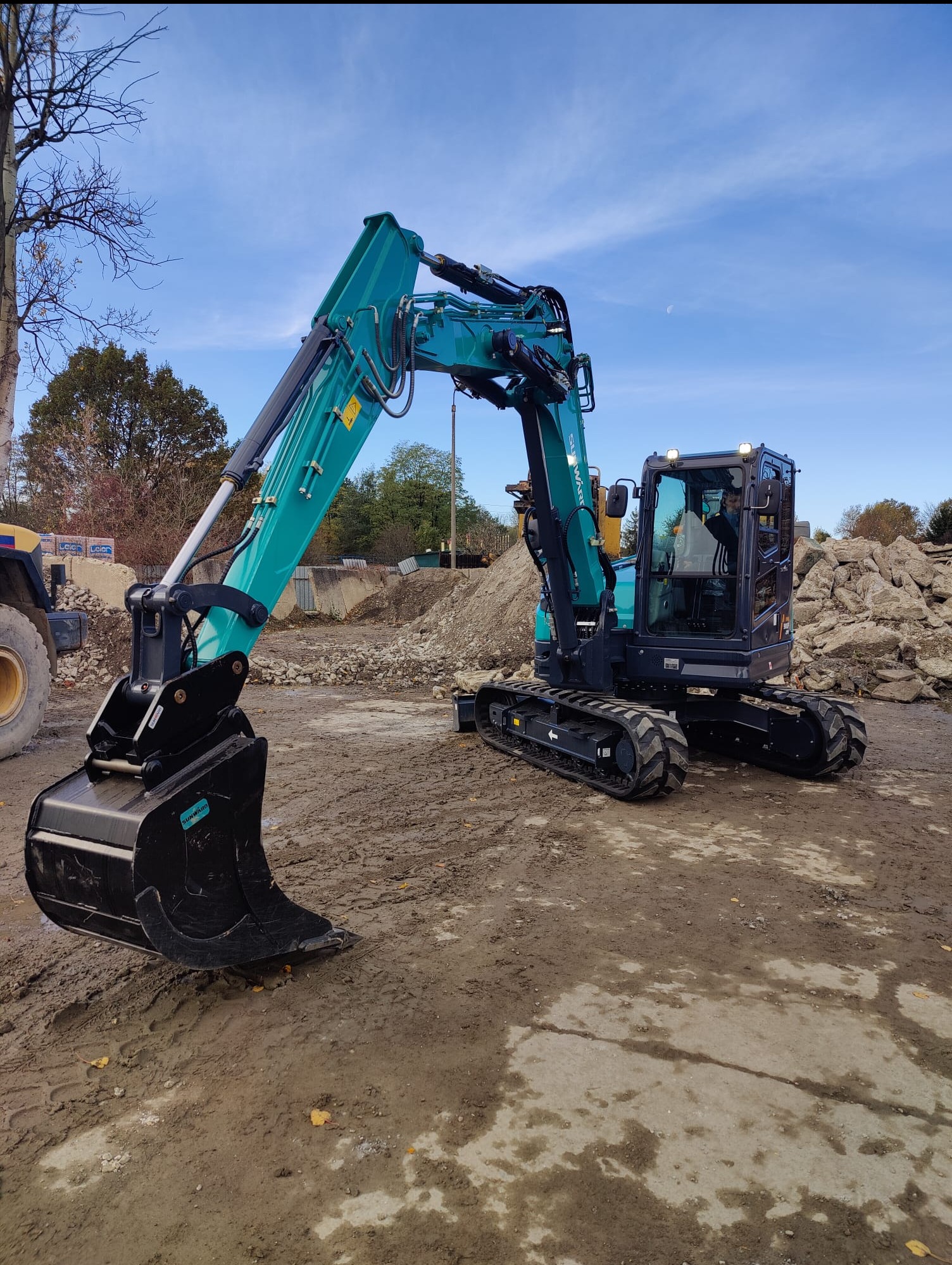 Niebiesko-czarna minikoparka gąsienicowa marki Sunward, zaparkowana na placu budowy, z widocznym stosem gruzu w tle i fragmentem żółtej maszyny budowlanej po lewej stronie.