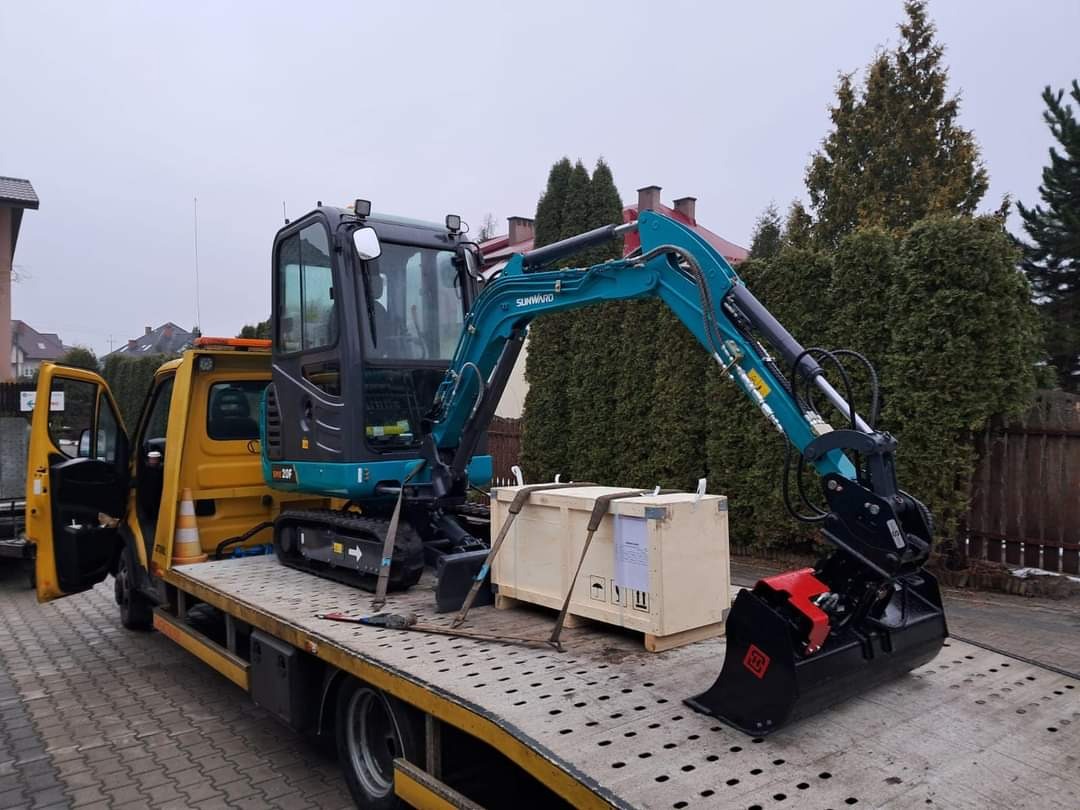 Niebiesko-czarna mini koparka Sunward 20F, zabezpieczona pasami transportowymi, na platformie żółtej lawety, obok skrzynia transportowa, w tle zabudowa jednorodzinna.