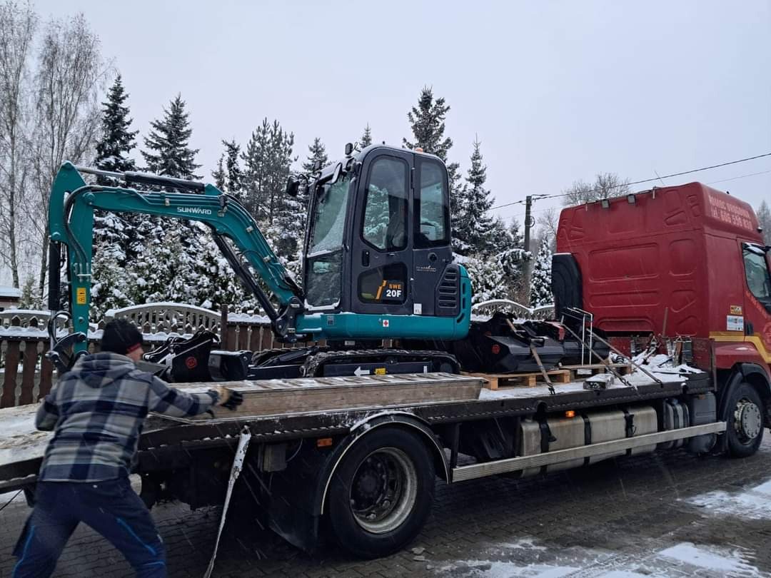 Niebiesko-szara mini koparka Sunward SWE 20F zabezpieczona na platformie czerwonej ciężarówki transportowej; w tle ośnieżone drzewa i budynki.