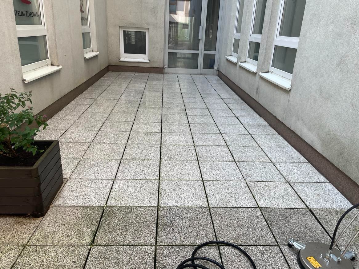Fragment kostki brukowej przed budynkiem, część wyczyszczona, widoczne różnice w kolorze, w rogu donica z zielenią i urządzenie do mycia ciśnieniowego.