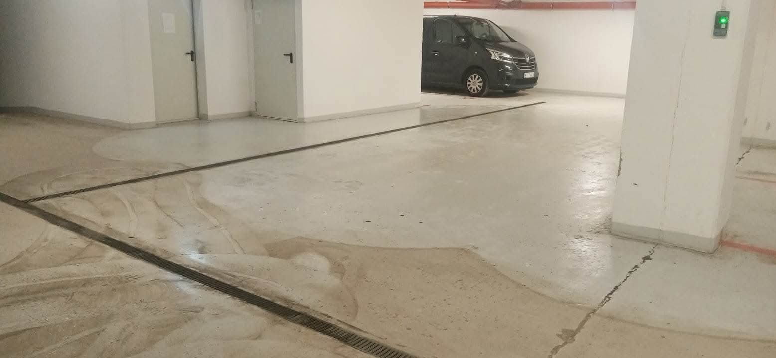 Wnętrze parkingu podziemnego z widocznymi śladami mycia posadzki, w tle zaparkowany czarny van.