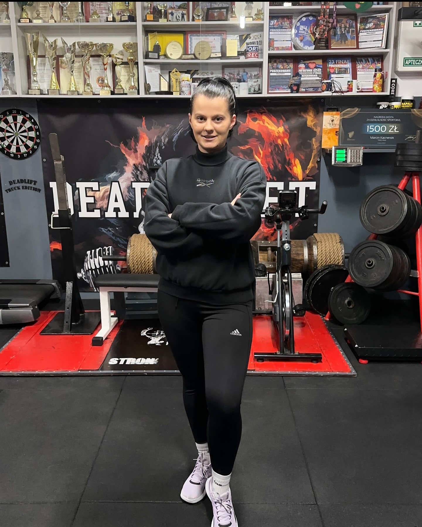 Kobieta w czarnym dresie stoi na tle siłowni z trofeami, sprzętem do ćwiczeń siłowych i dartem na ścianie.