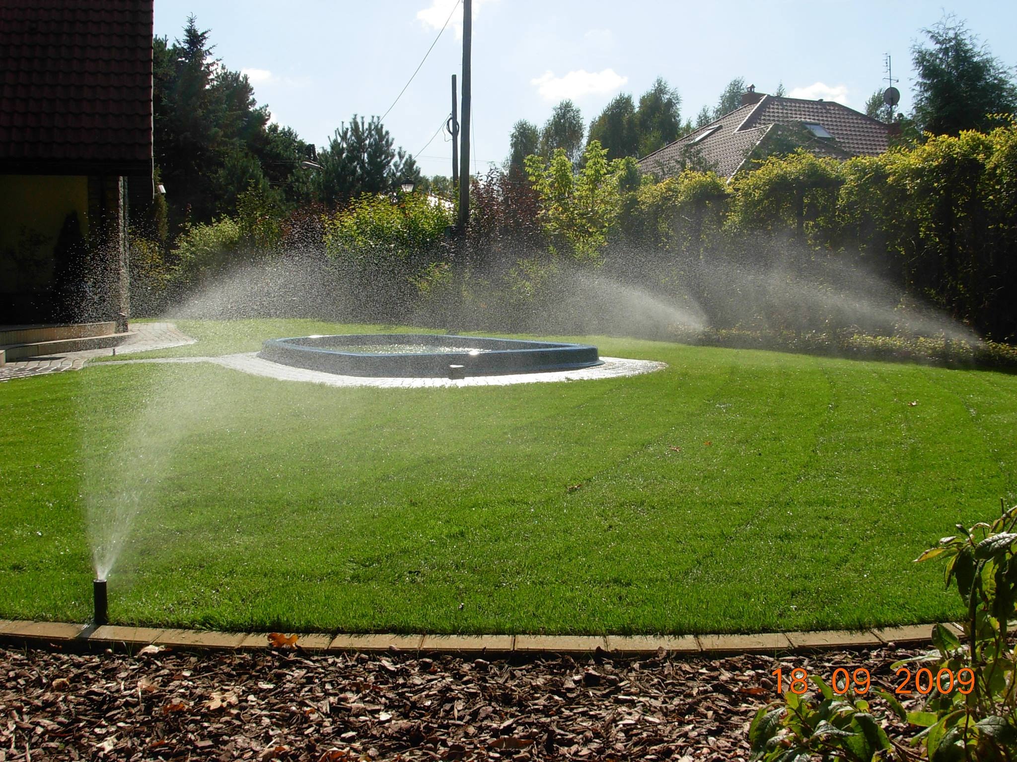 Automatyczne nawadnianie trawnika zraszaczami rotacyjnymi, w tle mały zbiornik wodny, wokół drzewa i dom.