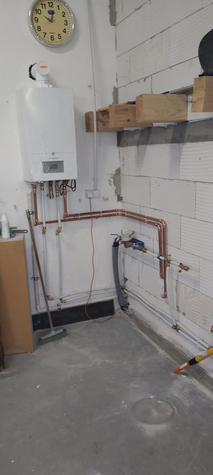 Świeżo zamontowany, biały kocioł gazowy z miedzianą instalacją, widoczny zegar ścienny i fragment ściany z betonu komórkowego w surowym wnętrzu.