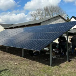 Instalacja paneli fotowoltaicznych na konstrukcji wsporczej, widoczni pracownicy podczas montażu, w tle budynek gospodarczy i błękitne niebo z chmurami.