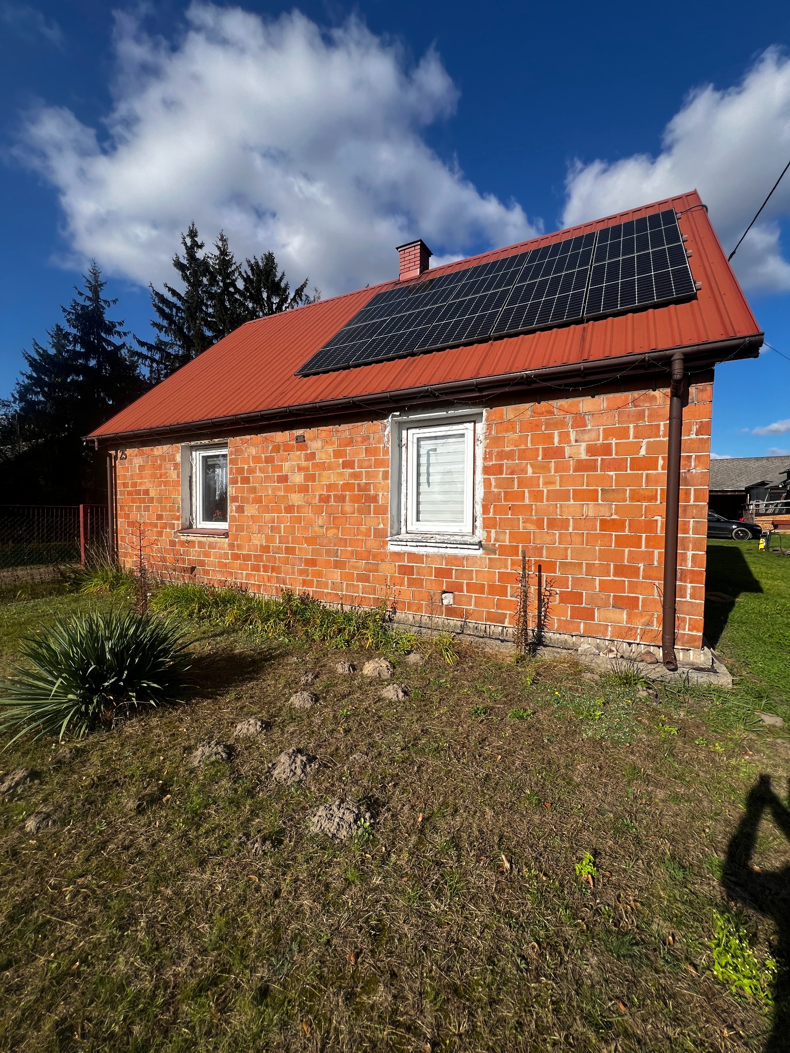 Niski, ceglany dom z czerwonym dachem, na którym zamontowano panele słoneczne; widok z poziomu gruntu na tle błękitnego nieba z chmurami.