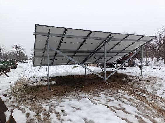 Montaż paneli słonecznych w zimowej scenerii w okolicach Warszawy, widok od spodu na stalową konstrukcję wsporczą i moduły fotowoltaiczne w trakcie instalacji na zasnieżonym terenie.