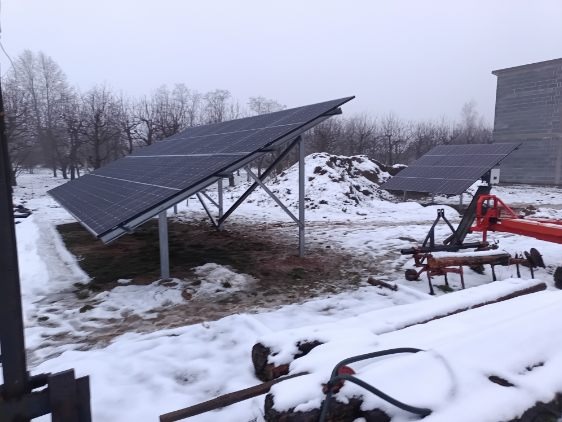 Dwa panele słoneczne na metalowych konstrukcjach w zimowym krajobrazie, z resztkami śniegu na ziemi i sprzętem rolniczym w tle.