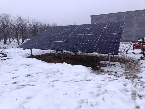 Panel fotowoltaiczny na metalowych podporach, zainstalowany na zaśnieżonym terenie w pobliżu drzew bez liści i budynku gospodarczego w pochmurny dzień.