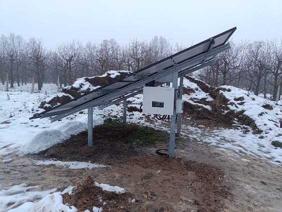 Panel słoneczny zamontowany na metalowej konstrukcji na zaśnieżonym terenie z drzewami w tle, widoczny falownik zamontowany na konstrukcji.