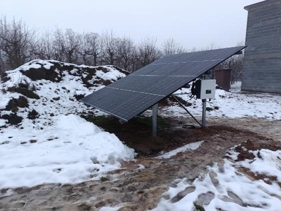 Panel słoneczny ustawiony na metalowych podporach na zasypanym śniegiem podwórku, z budynkiem z szarej cegły w tle i drzewami bez liści.