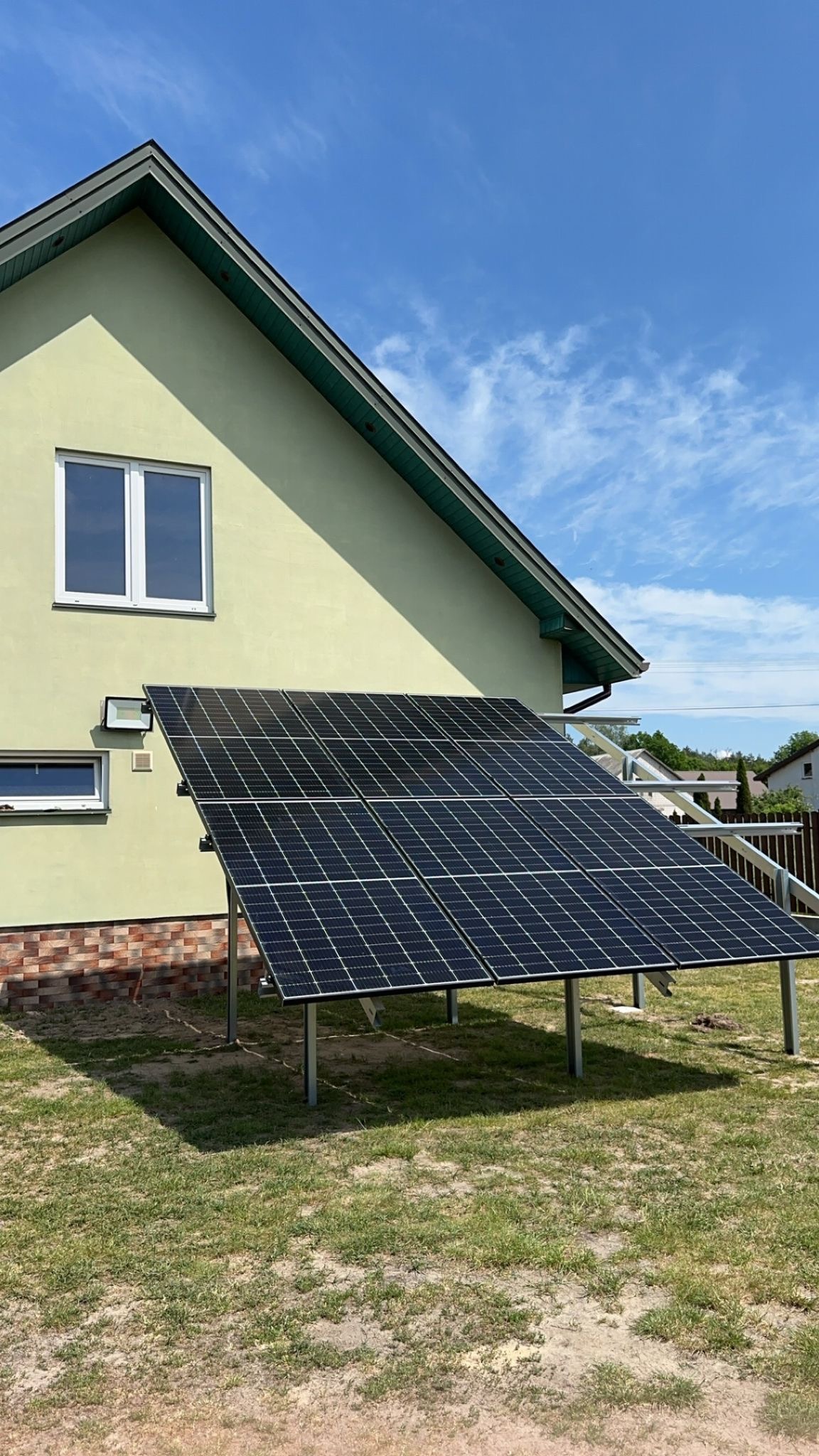 Panele słoneczne zamontowane na metalowych wspornikach obok domu z oknami, na tle błękitnego nieba z chmurami.