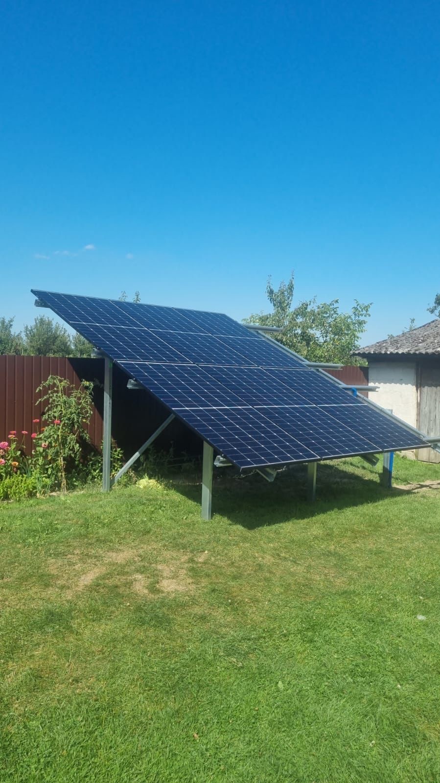 Nisko ustawiony panel słoneczny na metalowej konstrukcji w ogrodzie, z brązowym ogrodzeniem i kwiatami w tle, pod błękitnym niebem.
