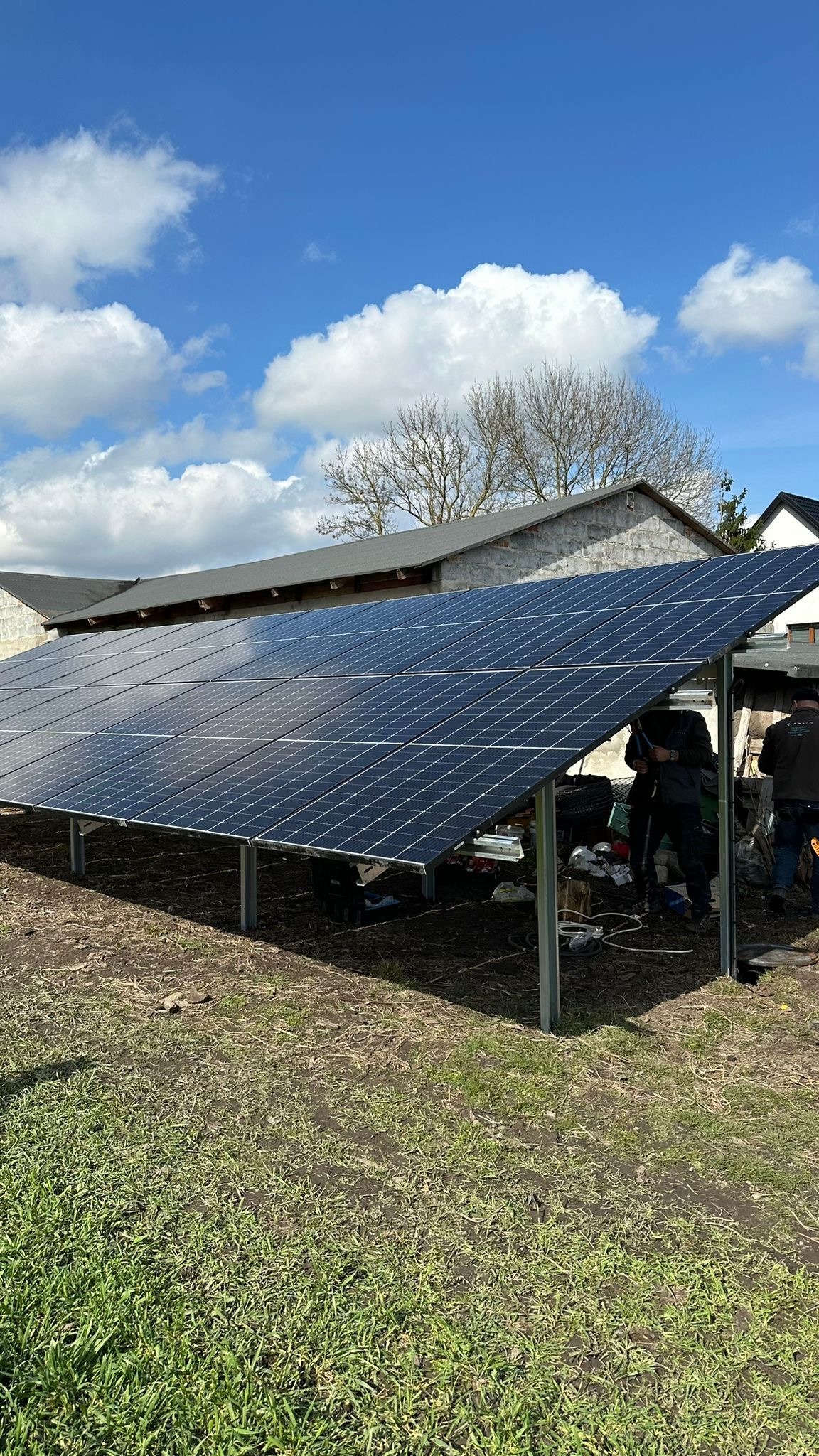 Instalacja paneli fotowoltaicznych na konstrukcji wsporczej, widoczni pracownicy podczas montażu, w tle budynek gospodarczy i błękitne niebo z chmurami.
