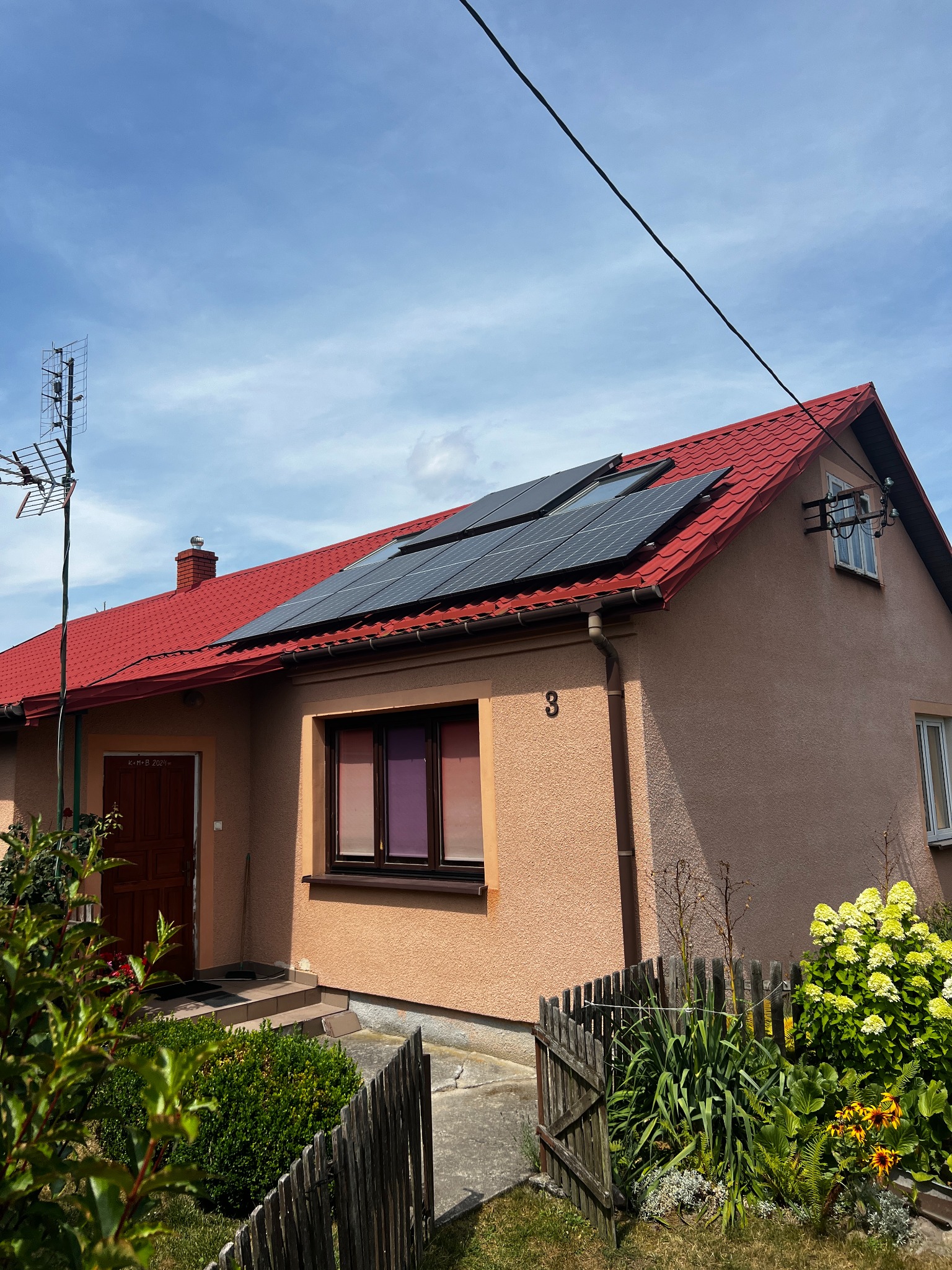 Dom jednorodzinny z czerwonym dachem i panelami słonecznymi, z numerem 3 na elewacji, otoczony zielenią i drewnianym płotem.
