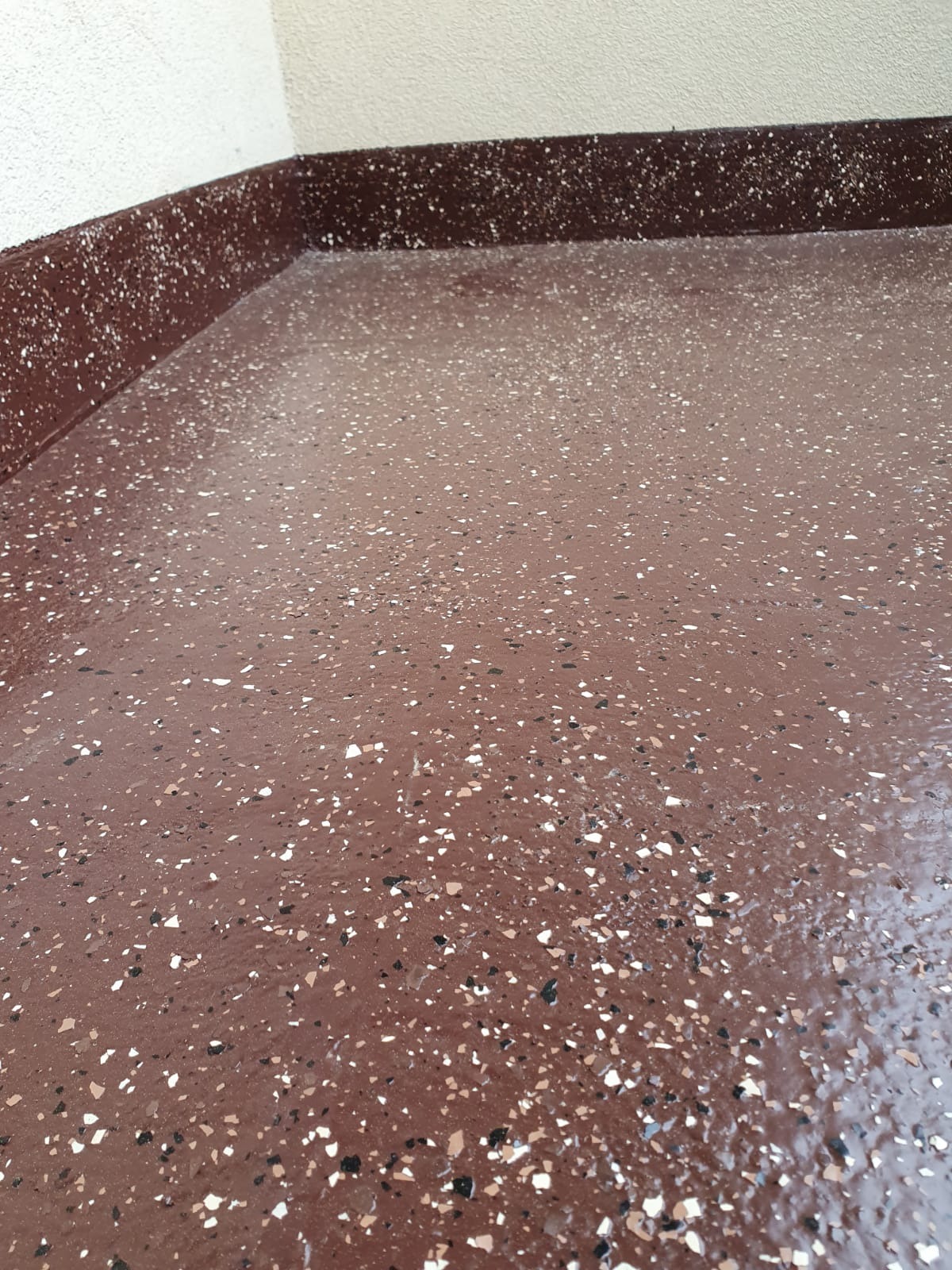 Błyszcząca, brązowa posadzka żywiczna z kolorowymi drobinkami, tworząca efekt lastryko, widoczna przy białej ścianie.