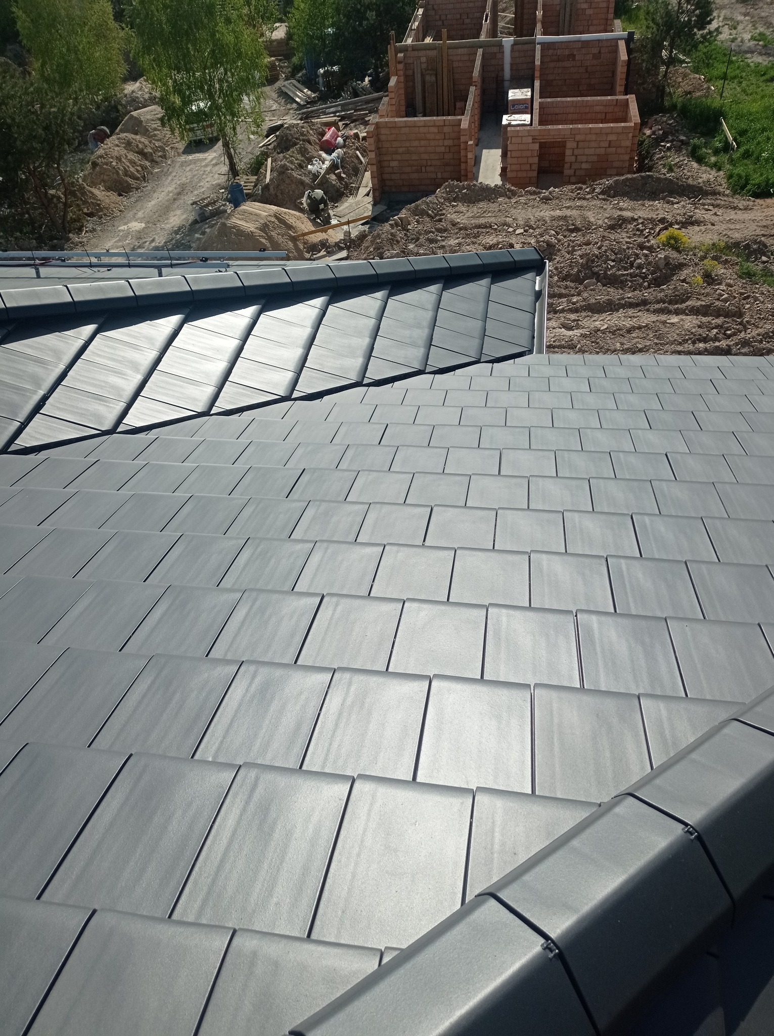 Nowy dach pokryty szarą dachówką ceramiczną, widok z góry na ułożone rzędy, w tle plac budowy z surową konstrukcją budynku z cegły.