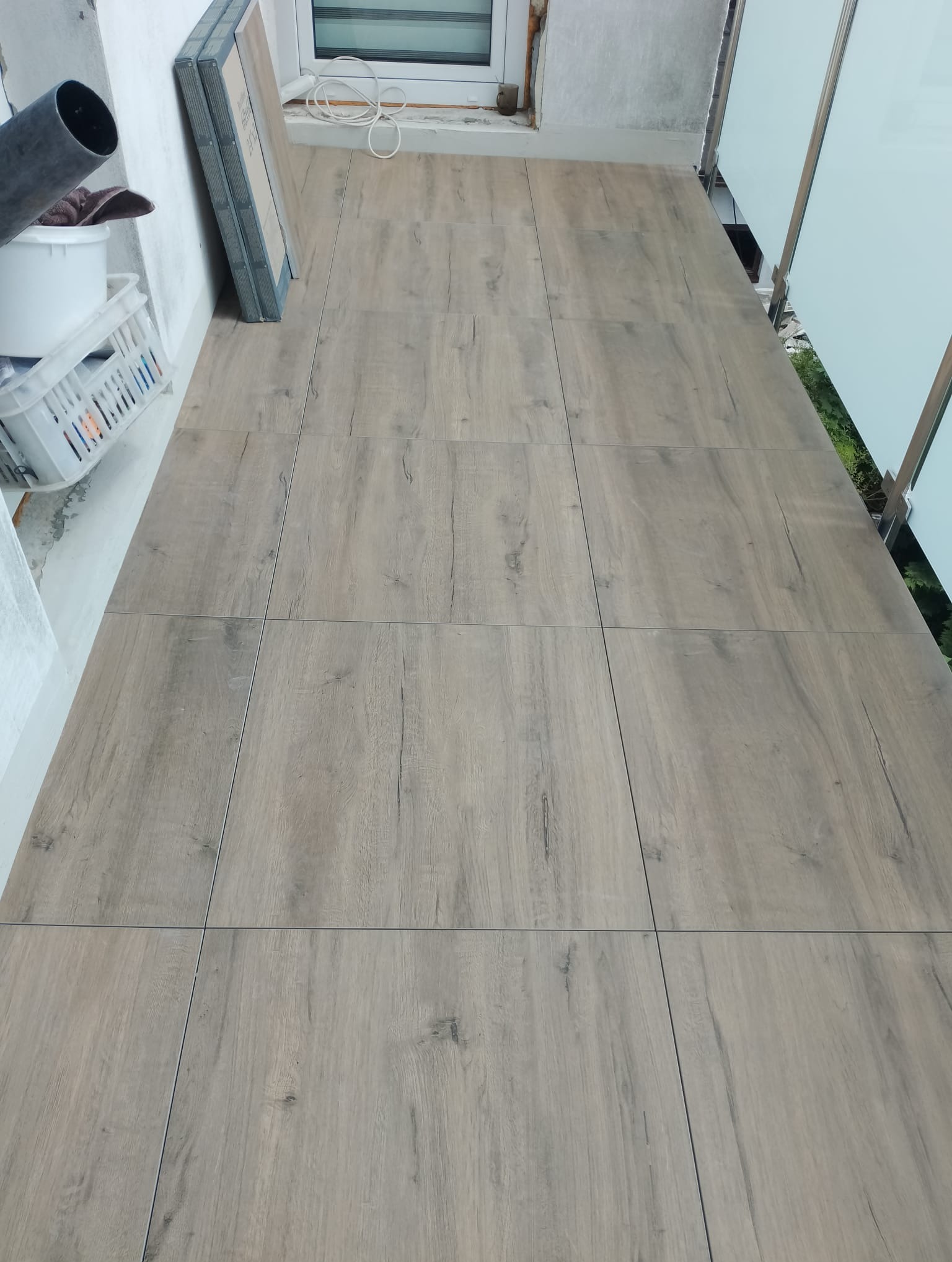 Ułożona podłoga z płytek imitujących drewno na balkonie, widoczne szklane balustrady i materiały budowlane.
