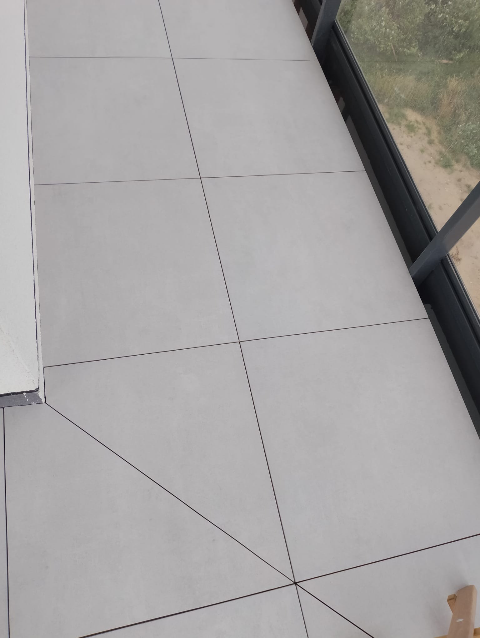 Szare płytki ceramiczne na balkonie lub tarasie, widok z góry, z widoczną ramą okienną i fragmentem krajobrazu za oknem.