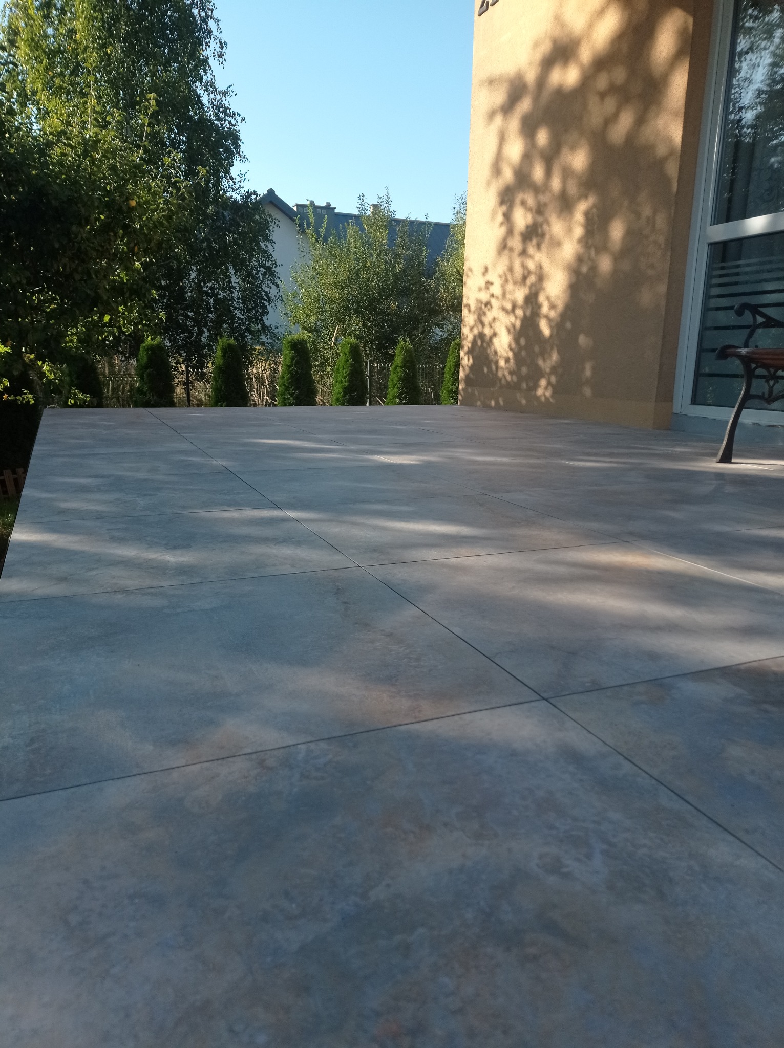 Wyłożony szarymi płytami taras przed budynkiem, widoczne drzewa i krzewy w tle, cień drzewa na ścianie budynku.