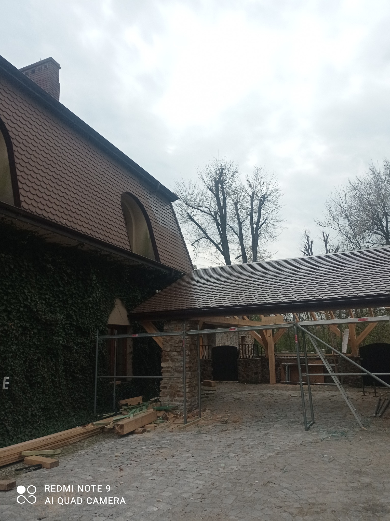 Budynek z dachem pokrytym brązową dachówką w łuskę, częściowo porośnięty bluszczem, z dobudowaną drewnianą konstrukcją z kamiennymi filarami, otoczoną rusztowaniami i ułożonymi belkami drewna...