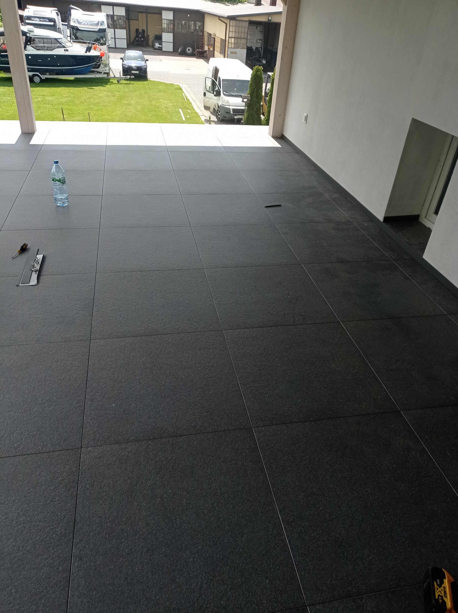 Ciemnoszara, wyłożona płytkami podłoga na zewnątrz budynku, z widoczną butelką wody, narzędziami i fragmentem zielonego trawnika z pojazdami w tle.