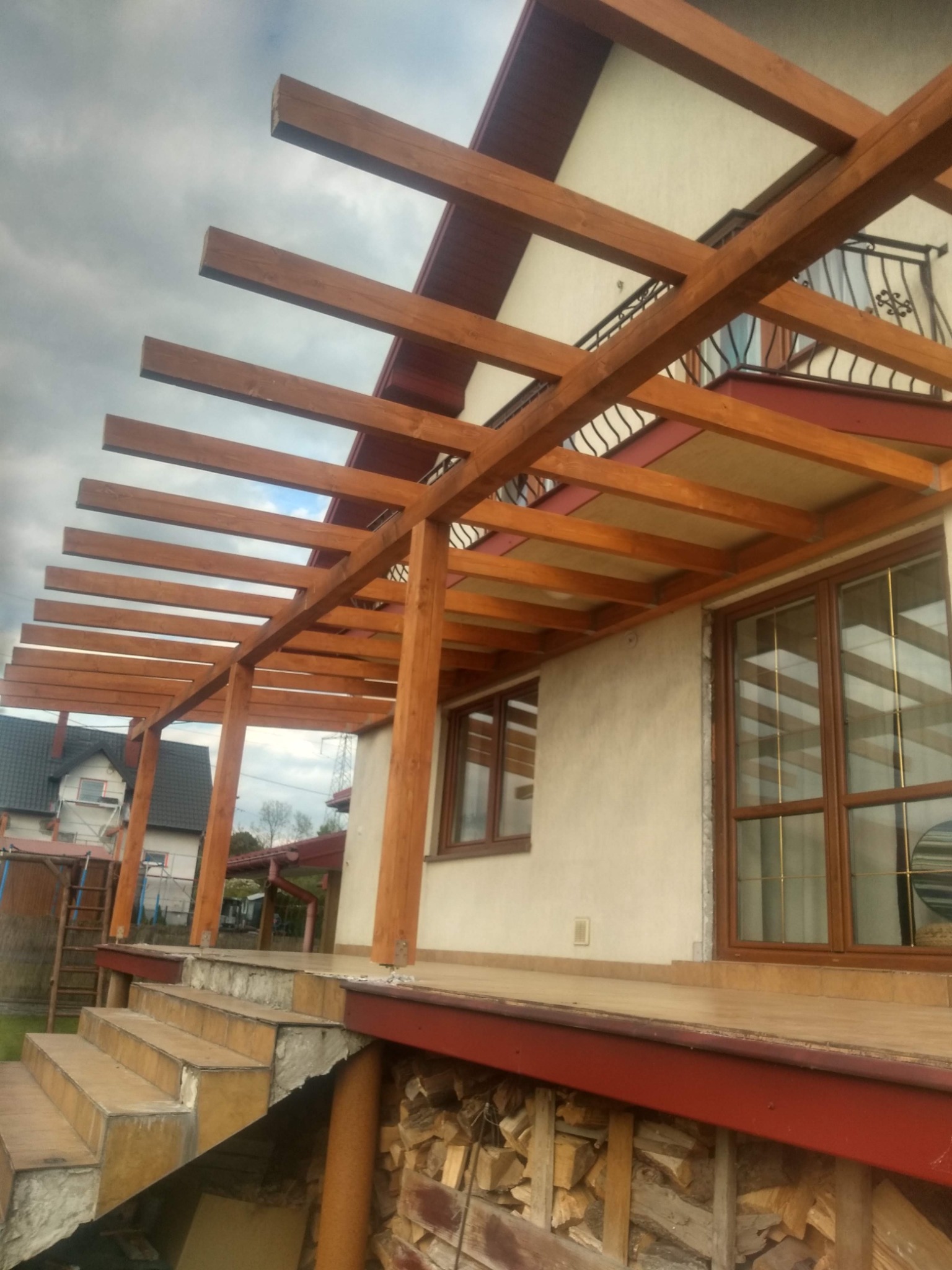 Drewniana konstrukcja z belek nad tarasem, częściowo zadaszona, z widocznymi schodami wejściowymi i składowanym drewnem pod spodem.