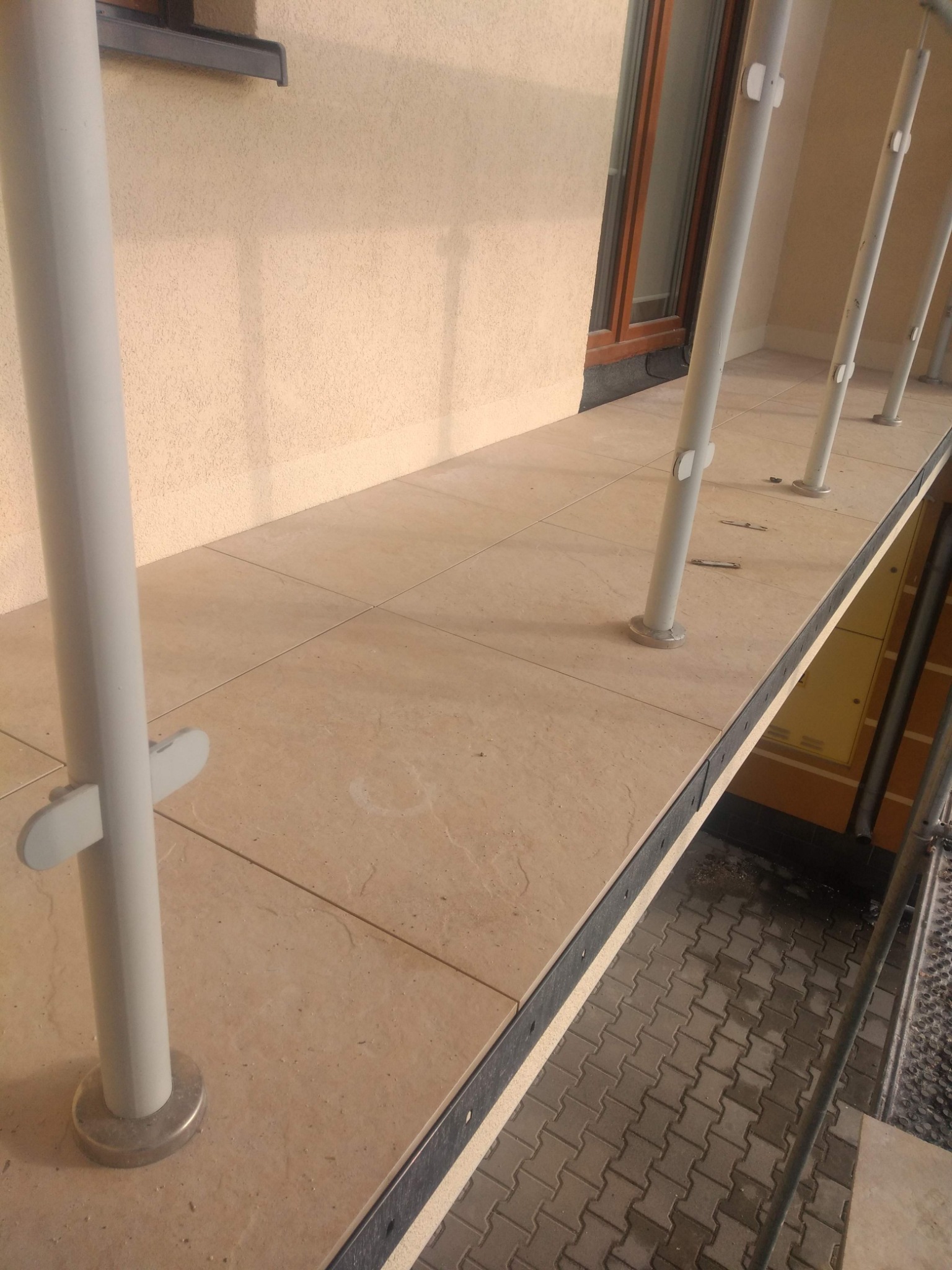 Wykończony balkon z beżowymi płytkami, balustradą z jasnych, pionowych słupków i widokiem na kostkę brukową poniżej.