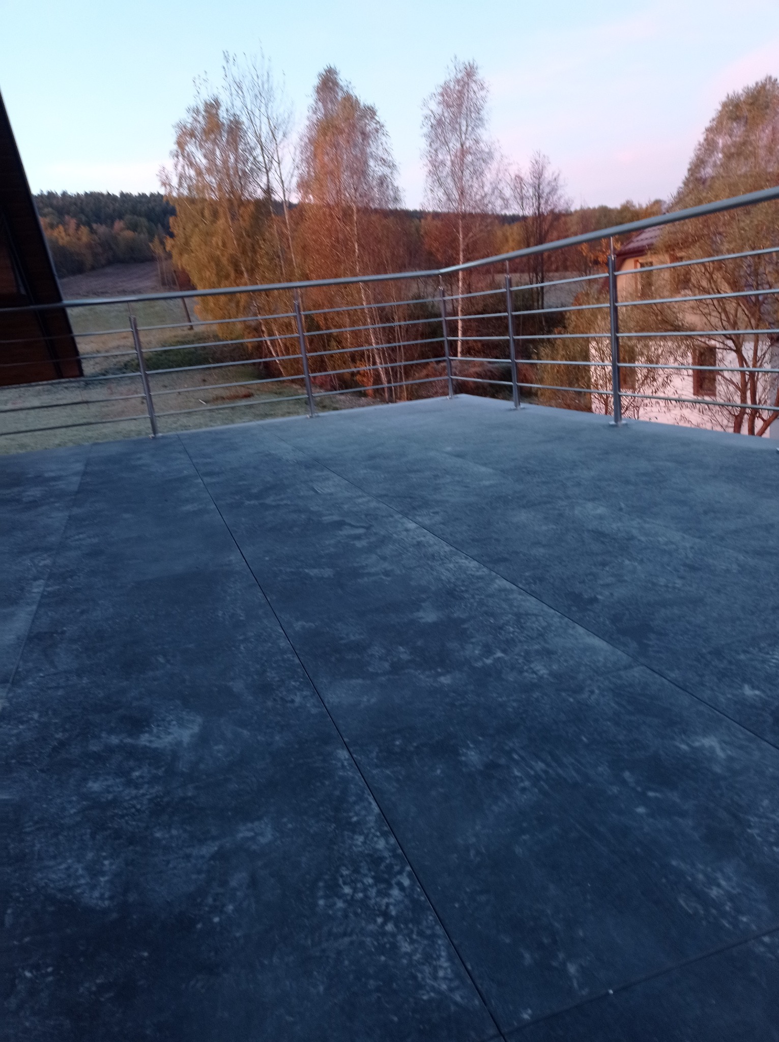 Wykończony taras z ciemnych płyt na zewnątrz, z metalową balustradą i widokiem na drzewa w tle o zmierzchu.