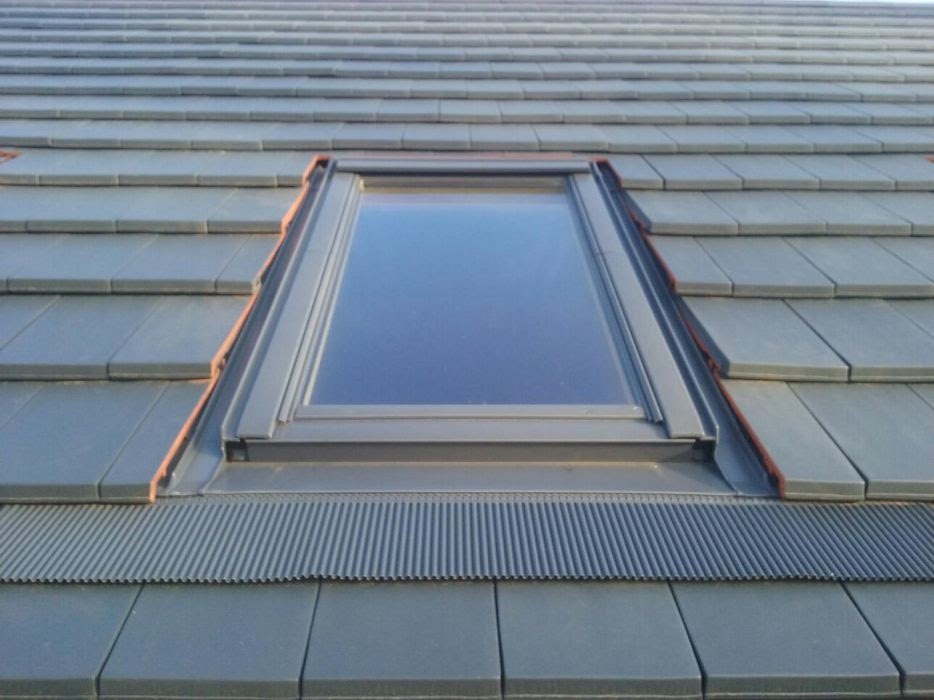 Szary dach pokryty dachówką z zamontowanym oknem dachowym z czarną ramą i elementami uszczelniającymi w kolorze ceglastym.