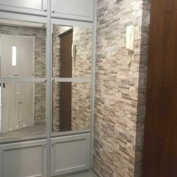 Marek Gdański - Szara zabudowa szafy z lustrami w przedpokoju, ściana wykończona tapetą imitującą kamień, widoczny domofon i gniazdko elektryczne.