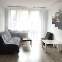 Marek Gdański - Jasne wnętrze pokoju dziennego z panelami podłogowymi, szarą kanapą przykrytą pledem, dwoma stolikami kawowymi i obrazem z motywem afrykańskim na ścianie.