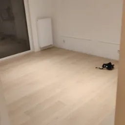 Jasne panele podłogowe w pokoju z białymi ścianami, grzejnikiem i czarną maską leżącą na podłodze, widoczny kabel elektryczny przy listwie przypodłogowej.