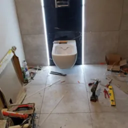 Montaż podwieszanej toalety na tle czarnej płyty z marmurowym wzorem, podświetlonej po bokach, widoczne narzędzia glazurnicze i płyty podłogowe oraz częściowo położona glazura ścienna.