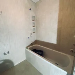 Wnętrze łazienki w trakcie prac: ułożone płytki w odcieniach beżu z pionowymi żłobieniami na ścianie, zabudowana wanna z widocznymi narzędziami w środku, przygotowane wnęki w ścianie.