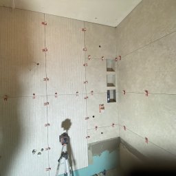 Marek Gdański - Ściany łazienki w trakcie układania płytek, widoczne poziomice laserowe, krzyżyki dystansowe i częściowo położone płytki w dwóch różnych wzorach.