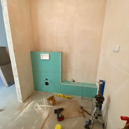 Marek Gdański - Montaż stelaża podtynkowego z zielonej płyty gipsowo-kartonowej w surowym wnętrzu łazienki, widoczne narzędzia i poziomica na podłodze, ściany w trakcie szpachlowania.