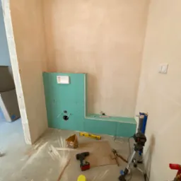 Montaż stelaża podtynkowego z zielonej płyty gipsowo-kartonowej w surowym wnętrzu łazienki, widoczne narzędzia i poziomica na podłodze, ściany w trakcie szpachlowania.