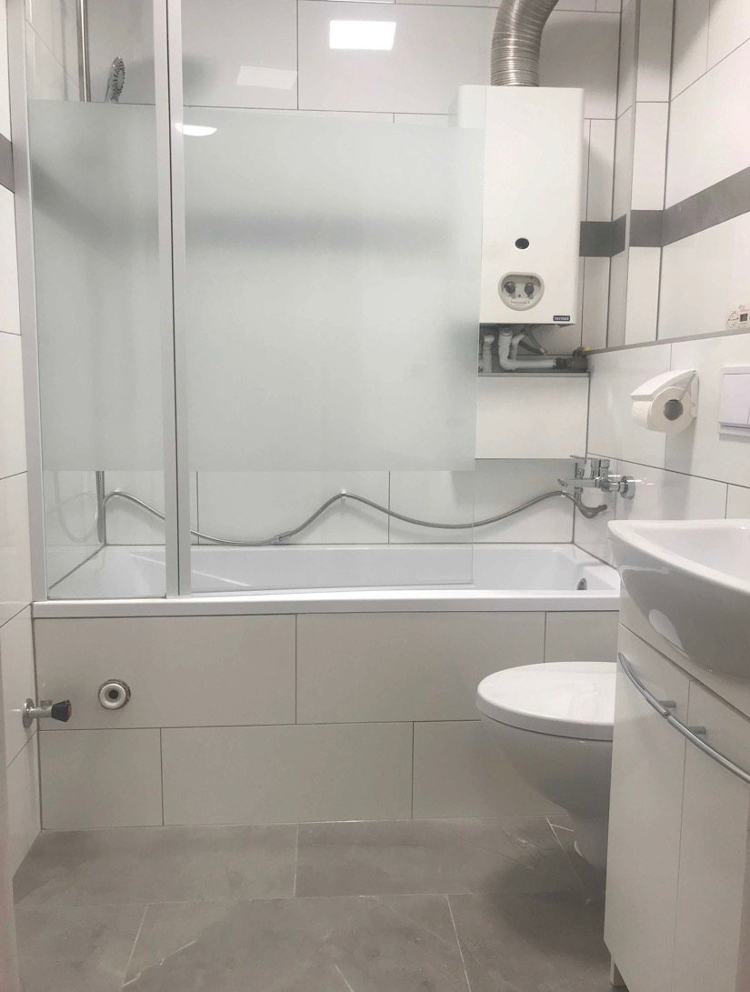 Biała łazienka z kabiną prysznicową z matową szybą, piecem gazowym nad wanną, umywalką z szafką i toaletą, wykończona płytkami w odcieniach bieli i szarości.