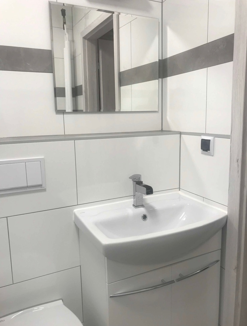 Biała umywalka z szafką w rogu łazienki, chromowana bateria z kaskadowym strumieniem wody, białe płytki z szarym pasem dekoracyjnym, lustro z szafkami w tle.