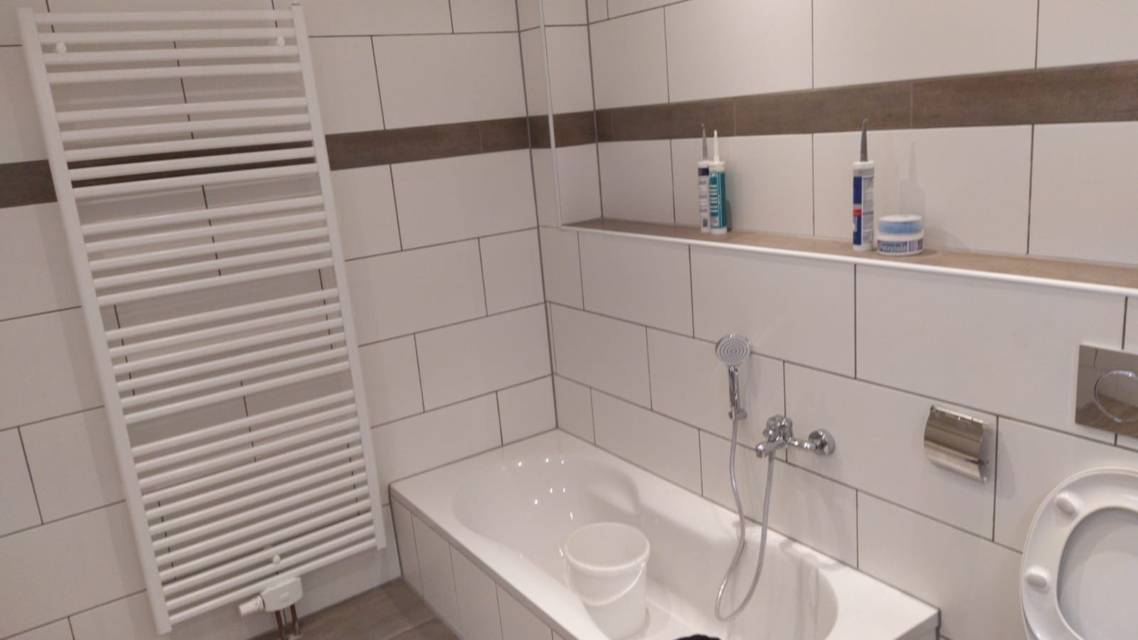 Wykończona łazienka z białymi płytkami, grzejnikiem drabinkowym, wanną, armaturą i półką z materiałami budowlanymi.