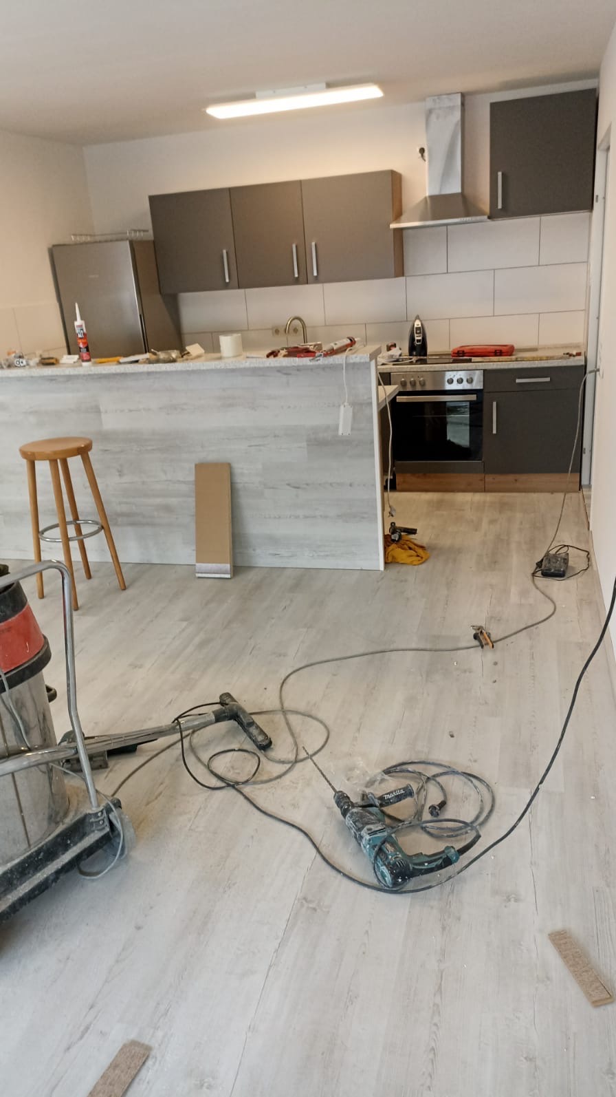 Wnętrze kuchni w trakcie remontu: widoczne narzędzia, odkurzacz przemysłowy, nieukończona podłoga z paneli, częściowo zamontowane szafki kuchenne w odcieniach szarości i białe płytki na ścianie.