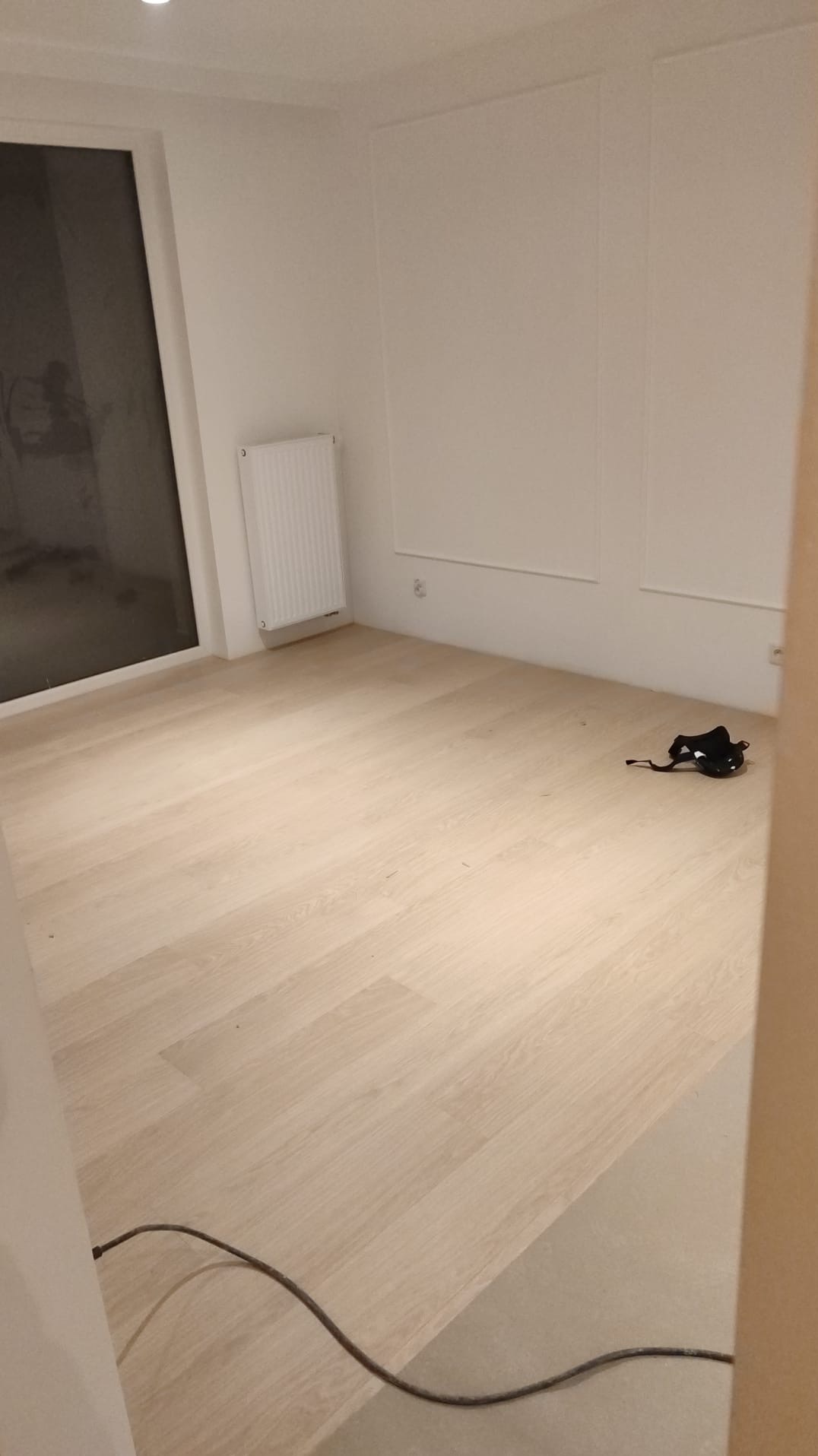 Jasne panele podłogowe w pokoju z białymi ścianami, grzejnikiem i czarną maską leżącą na podłodze, widoczny kabel elektryczny przy listwie przypodłogowej.